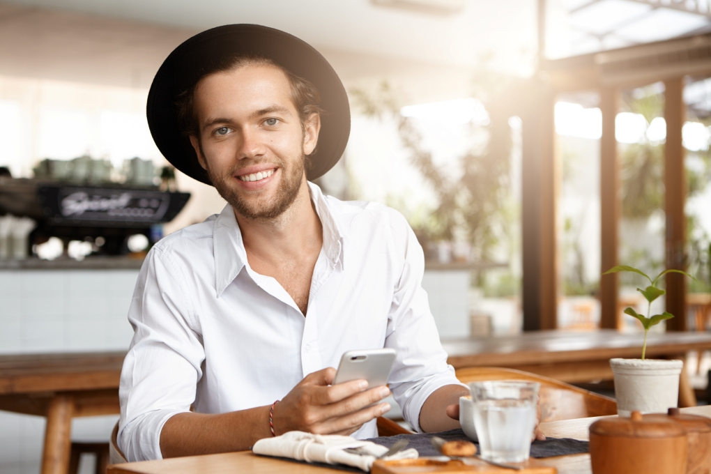 Attractive happy young bearded businessman in trendy hat texting messages via social networks and browsing internet, using free wifi on his electronic device during coffee break at restaurant Attractive happy young bearded businessman in trendy hat texting messages via social networks and browsing internet, using free wifi on his electronic device during coffee break at restaurant